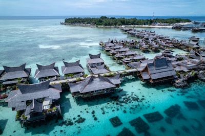 A cluster of traditional wooden houses on stilts located over clear turquoise water. The structures have pointed roofs and are connected by narrow walkways. In the background, a lush green island is visible, and the overall setting suggests a scenic tropical resort.