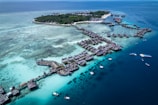 Aerial view of a tropical island resort featuring numerous overwater bungalows surrounded by turquoise and deep blue waters. The island is lush with green vegetation and surrounded by a vibrant reef. Several boats are visible in the water, highlighting the area's idyllic and serene setting.