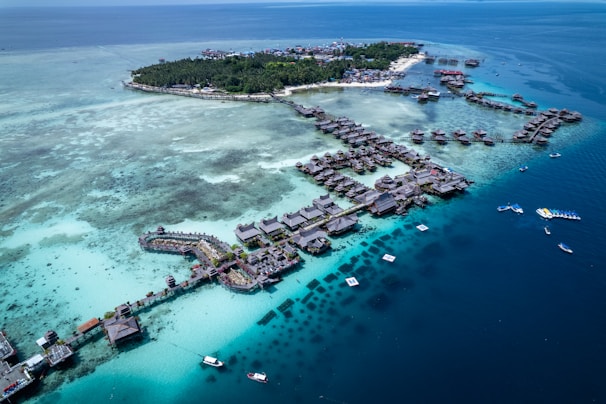 Aerial view of a tropical island resort featuring numerous overwater bungalows surrounded by turquoise and deep blue waters. The island is lush with green vegetation and surrounded by a vibrant reef. Several boats are visible in the water, highlighting the area's idyllic and serene setting.
