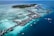 Aerial view of a tropical island resort featuring numerous overwater bungalows surrounded by turquoise and deep blue waters. The island is lush with green vegetation and surrounded by a vibrant reef. Several boats are visible in the water, highlighting the area's idyllic and serene setting.