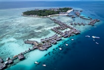Aerial view of a tropical island resort featuring numerous overwater bungalows surrounded by turquoise and deep blue waters. The island is lush with green vegetation and surrounded by a vibrant reef. Several boats are visible in the water, highlighting the area's idyllic and serene setting.