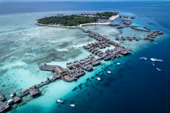 Aerial view of a tropical island resort featuring numerous overwater bungalows surrounded by turquoise and deep blue waters. The island is lush with green vegetation and surrounded by a vibrant reef. Several boats are visible in the water, highlighting the area's idyllic and serene setting.