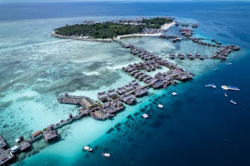 Aerial view of a tropical island resort featuring numerous overwater bungalows surrounded by turquoise and deep blue waters. The island is lush with green vegetation and surrounded by a vibrant reef. Several boats are visible in the water, highlighting the area's idyllic and serene setting.