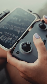 A person is holding a drone controller in one hand and a smartphone in the other. The controller has joysticks and buttons, while the smartphone screen displays a blurred interface. The person's nails are painted purple.