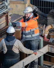 Two construction workers, wearing safety helmets and protective clothing, are standing at a construction site. One is wearing an orange high-visibility jacket, smiling, and gesturing with his arms. The background includes various construction materials and equipment.