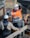 Two construction workers, wearing safety helmets and protective clothing, are standing at a construction site. One is wearing an orange high-visibility jacket, smiling, and gesturing with his arms. The background includes various construction materials and equipment.