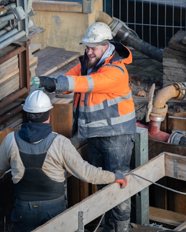 Two construction workers, wearing safety helmets and protective clothing, are standing at a construction site. One is wearing an orange high-visibility jacket, smiling, and gesturing with his arms. The background includes various construction materials and equipment.