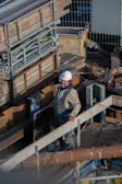 A construction worker wearing a white hard hat and overalls is standing on a wooden scaffolding surrounded by building materials and pipes. The worker appears to be engaged in some aspect of construction work, possibly handling a large plank or piece of equipment.
