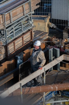 A construction worker wearing a white hard hat and overalls is standing on a wooden scaffolding surrounded by building materials and pipes. The worker appears to be engaged in some aspect of construction work, possibly handling a large plank or piece of equipment.