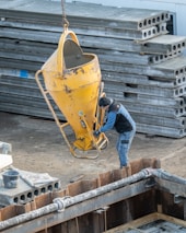 A construction site scene with a worker handling a large yellow concrete funnel suspended by a chain. Several stacks of precast concrete slabs are visible in the background. The ground is covered with construction materials and equipment.