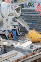 A construction site with a concrete mixer pouring concrete into a large yellow container. Several metal structures and concrete slabs are scattered around. A worker wearing a blue jacket and dark pants is standing next to the mixer, overseeing the process.
