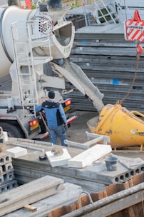 A construction site with a concrete mixer pouring concrete into a large yellow container. Several metal structures and concrete slabs are scattered around. A worker wearing a blue jacket and dark pants is standing next to the mixer, overseeing the process.