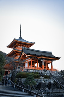 A traditional Japanese temple with intricate wooden architecture, featuring bright orange accents and a multi-tiered pagoda roof. The temple is situated on a stone platform with a staircase leading up to it, surrounded by well-manicured greenery and stone lanterns. The setting is serene, with a clear blue sky overhead.