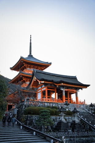 A traditional Japanese temple with intricate wooden architecture, featuring bright orange accents and a multi-tiered pagoda roof. The temple is situated on a stone platform with a staircase leading up to it, surrounded by well-manicured greenery and stone lanterns. The setting is serene, with a clear blue sky overhead.