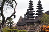 A series of intricately designed towers with tiered roofs, typical of traditional Balinese architecture, are surrounded by lush greenery and tropical plants. The atmosphere is serene and culturally rich, with the ancient structure standing out against a somewhat cloudy sky.
