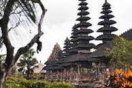 A series of intricately designed towers with tiered roofs, typical of traditional Balinese architecture, are surrounded by lush greenery and tropical plants. The atmosphere is serene and culturally rich, with the ancient structure standing out against a somewhat cloudy sky.
