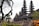 A series of intricately designed towers with tiered roofs, typical of traditional Balinese architecture, are surrounded by lush greenery and tropical plants. The atmosphere is serene and culturally rich, with the ancient structure standing out against a somewhat cloudy sky.