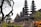 A series of intricately designed towers with tiered roofs, typical of traditional Balinese architecture, are surrounded by lush greenery and tropical plants. The atmosphere is serene and culturally rich, with the ancient structure standing out against a somewhat cloudy sky.