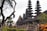 A series of intricately designed towers with tiered roofs, typical of traditional Balinese architecture, are surrounded by lush greenery and tropical plants. The atmosphere is serene and culturally rich, with the ancient structure standing out against a somewhat cloudy sky.