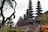 A series of intricately designed towers with tiered roofs, typical of traditional Balinese architecture, are surrounded by lush greenery and tropical plants. The atmosphere is serene and culturally rich, with the ancient structure standing out against a somewhat cloudy sky.