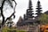 A series of intricately designed towers with tiered roofs, typical of traditional Balinese architecture, are surrounded by lush greenery and tropical plants. The atmosphere is serene and culturally rich, with the ancient structure standing out against a somewhat cloudy sky.