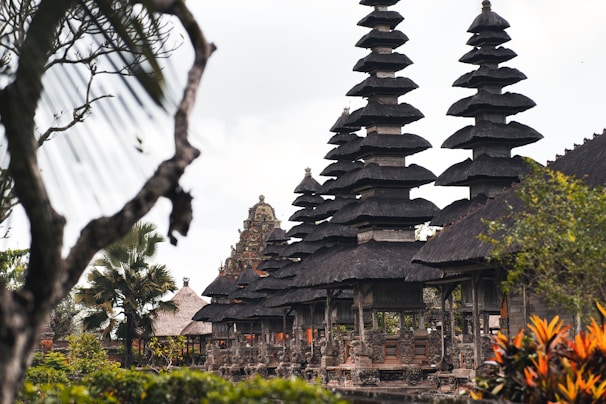 A series of intricately designed towers with tiered roofs, typical of traditional Balinese architecture, are surrounded by lush greenery and tropical plants. The atmosphere is serene and culturally rich, with the ancient structure standing out against a somewhat cloudy sky.