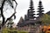 A series of intricately designed towers with tiered roofs, typical of traditional Balinese architecture, are surrounded by lush greenery and tropical plants. The atmosphere is serene and culturally rich, with the ancient structure standing out against a somewhat cloudy sky.