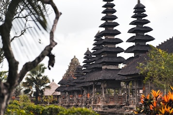 A series of intricately designed towers with tiered roofs, typical of traditional Balinese architecture, are surrounded by lush greenery and tropical plants. The atmosphere is serene and culturally rich, with the ancient structure standing out against a somewhat cloudy sky.