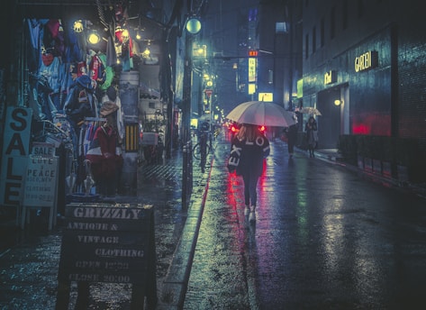 A dimly lit urban street scene at night in a rainy setting, with people walking under umbrellas. The street is lined with shops displaying vintage clothing and brightly colored garments. Neon lights and signs above cast colorful reflections on the wet pavement, adding to the moody ambiance.
