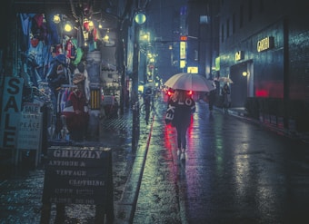 A dimly lit urban street scene at night in a rainy setting, with people walking under umbrellas. The street is lined with shops displaying vintage clothing and brightly colored garments. Neon lights and signs above cast colorful reflections on the wet pavement, adding to the moody ambiance.