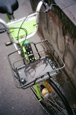 A bicycle with a green and white color scheme is parked against a wet stone surface. The bicycle features a front basket attached over the wheel, which is slick with rainwater. The seat is black, and the tires are thin. A printed sign with the words 'Yerevan Ride' is partially visible on the bike frame, suggesting a bike-sharing service.