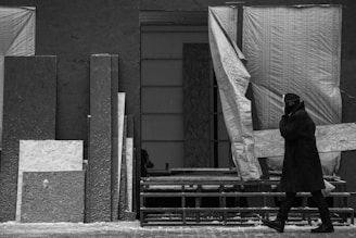 A person dressed in a heavy coat, hat, and gloves walks past a construction site or workshop. The background features stacked wooden and metal panels along with large draped fabrics, creating a textured and industrial setting. The scene is captured in black and white, emphasizing contrasts and shadows.