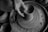 A black and white image depicting hands shaping clay on a pottery wheel. The texture of the wet clay is visible, with the artist's fingers skillfully molding a vessel form as the wheel spins. The surrounding area is scattered with clay remnants, emphasizing the hands-on process of pottery making.