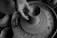 A black and white image depicting hands shaping clay on a pottery wheel. The texture of the wet clay is visible, with the artist's fingers skillfully molding a vessel form as the wheel spins. The surrounding area is scattered with clay remnants, emphasizing the hands-on process of pottery making.