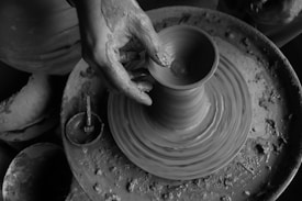 A black and white image depicting hands shaping clay on a pottery wheel. The texture of the wet clay is visible, with the artist's fingers skillfully molding a vessel form as the wheel spins. The surrounding area is scattered with clay remnants, emphasizing the hands-on process of pottery making.