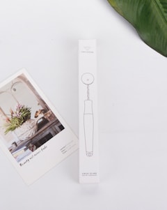 A minimalistic white background hosts a rectangular product box with line drawings and text in a non-English language. Adjacent to the box is an artistic card featuring a floral arrangement in a rustic setting with the text 'Beauty will never fade'. A green leaf partially frames the upper right corner.