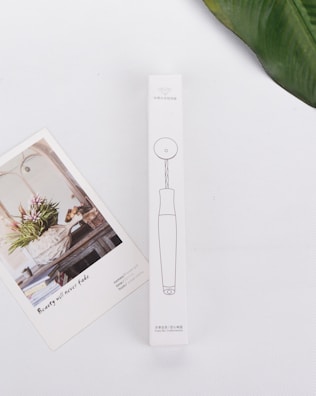 A minimalistic white background hosts a rectangular product box with line drawings and text in a non-English language. Adjacent to the box is an artistic card featuring a floral arrangement in a rustic setting with the text 'Beauty will never fade'. A green leaf partially frames the upper right corner.