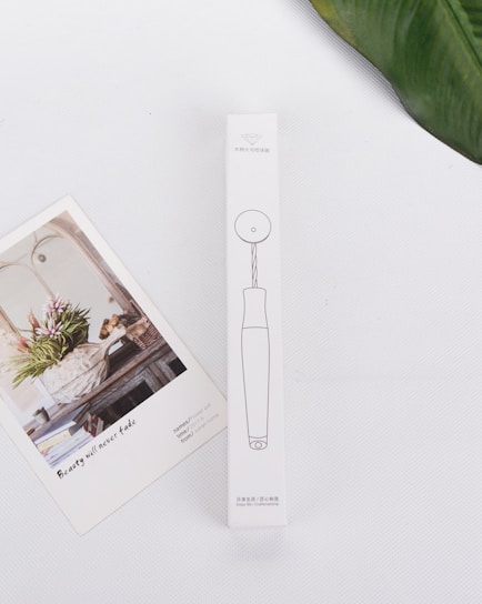A minimalistic white background hosts a rectangular product box with line drawings and text in a non-English language. Adjacent to the box is an artistic card featuring a floral arrangement in a rustic setting with the text 'Beauty will never fade'. A green leaf partially frames the upper right corner.