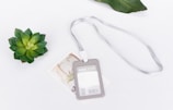 A plastic ID card holder with a silver lanyard lies on a white surface. Nearby, a small green succulent and a decorative card add a touch of nature and elegance.