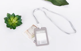 A plastic ID card holder with a silver lanyard lies on a white surface. Nearby, a small green succulent and a decorative card add a touch of nature and elegance.