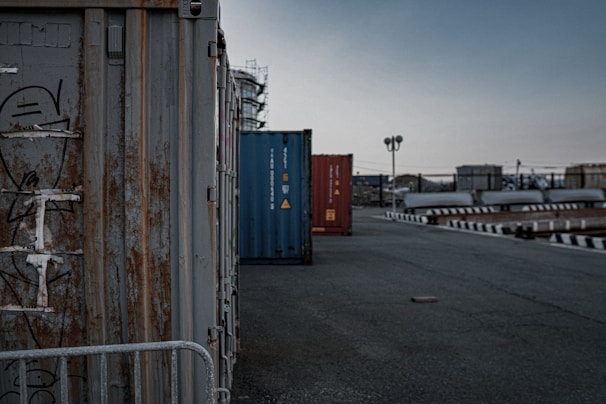A row of shipping containers is aligned along a deserted, industrial road. The closest container is rusted with some graffiti on it. The sky is overcast, contributing to a gloomy atmosphere. In the background, a few industrial structures and streetlights are visible.