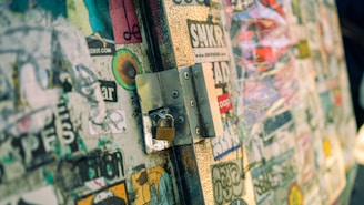 A close-up of a textured surface covered with various colorful stickers, featuring a metal latch and padlock. The stickers include a mix of text, logos, and abstract designs, some of which are weathered or peeling. The image has a shallow depth of field, with sharp focus on the latch and lock while the background appears blurred.