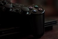A close-up of a black gaming controller resting on a gaming console. The controller features distinctive button icons, including a triangle, circle, cross, and square, each with unique colors. The lighting is dim, creating a moody and nostalgic atmosphere.