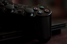 A close-up of a black gaming controller resting on a gaming console. The controller features distinctive button icons, including a triangle, circle, cross, and square, each with unique colors. The lighting is dim, creating a moody and nostalgic atmosphere.