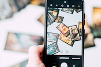 A hand is holding a smartphone, capturing an image of several tarot cards spread out in a scattered manner. The cards feature intricate and colorful designs, with various mystical and fantastical elements. The background is blurred, emphasizing the phone screen and the cards being displayed.