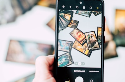 A hand is holding a smartphone, capturing an image of several tarot cards spread out in a scattered manner. The cards feature intricate and colorful designs, with various mystical and fantastical elements. The background is blurred, emphasizing the phone screen and the cards being displayed.