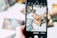 A hand is holding a smartphone, capturing an image of several tarot cards spread out in a scattered manner. The cards feature intricate and colorful designs, with various mystical and fantastical elements. The background is blurred, emphasizing the phone screen and the cards being displayed.