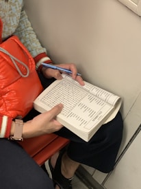 A person is sitting down, holding a word search puzzle book with one hand and using a blue pen to solve it. The individual is wearing a patterned sweater with red and white cuffs. A red quilted bag is resting on their lap, and they are seated on a red bench.
