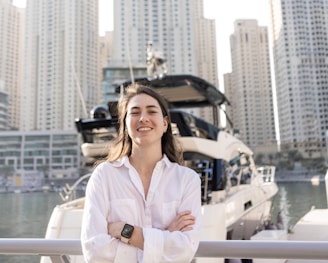 A person wearing a white shirt, smiling confidently with arms crossed, stands in front of a docked yacht. High-rise buildings form the background, reflecting a modern urban environment.