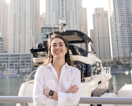 A person wearing a white shirt, smiling confidently with arms crossed, stands in front of a docked yacht. High-rise buildings form the background, reflecting a modern urban environment.